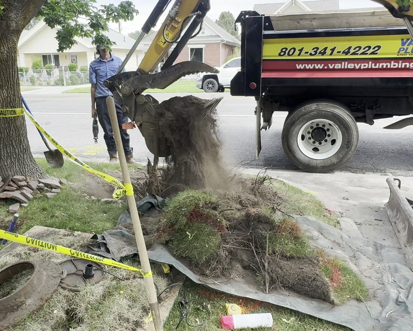 A Valley Plumbing technician and mini-excavator lifting a root ball out of a Utah front yard beside the company dump truck