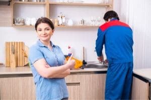 Plumber fixing leaky faucet, woman standing in kitchen giving thumbs up.