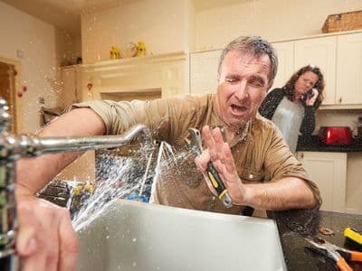 Man getting sprayed in the face by a broken sink pipe. Woman in background calling plumber.
